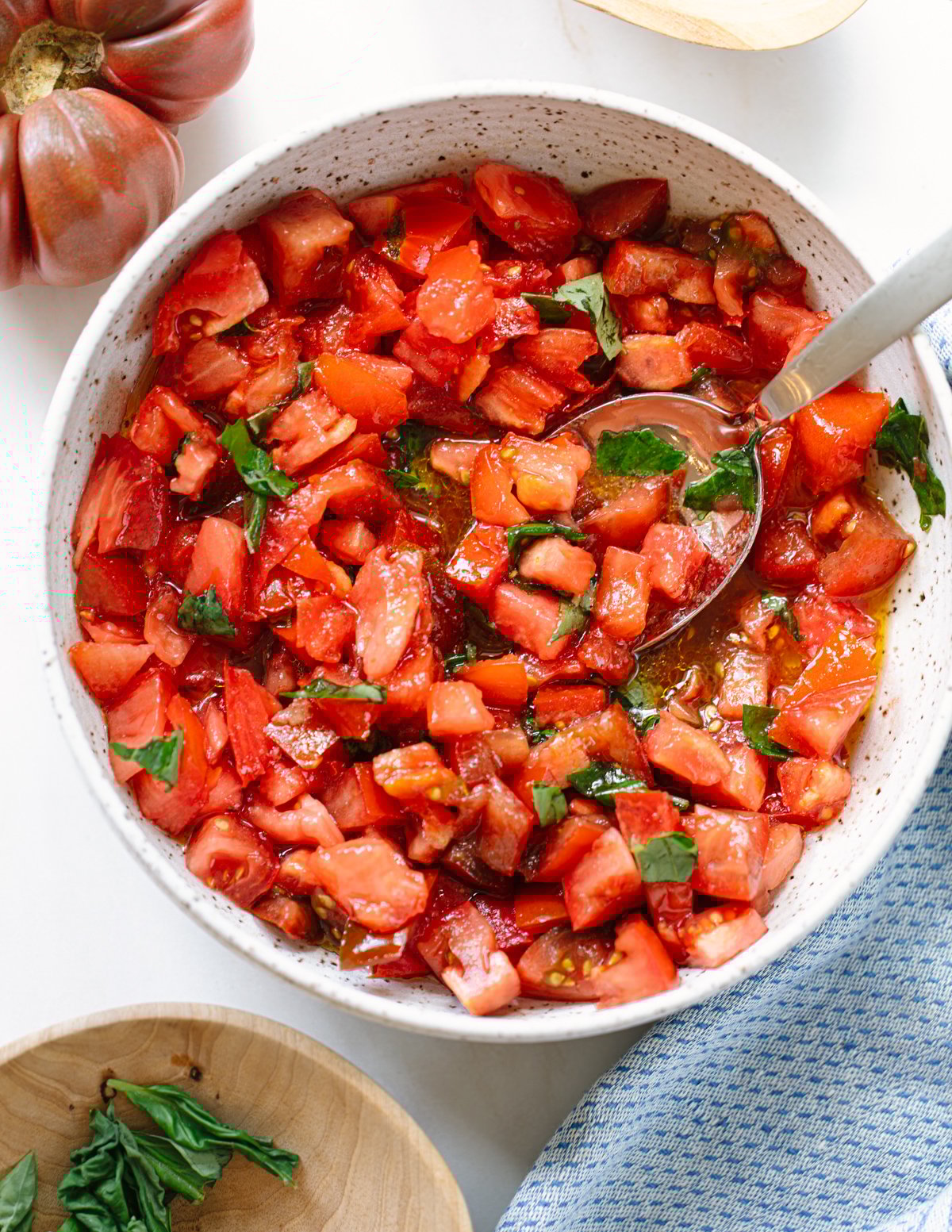 A bowl of freshly chopped tomatoes mixed with basil and olive oil, with a spoon inside the bowl. A whole tomato, basil leaves, and a blue cloth are nearby on a white surface.