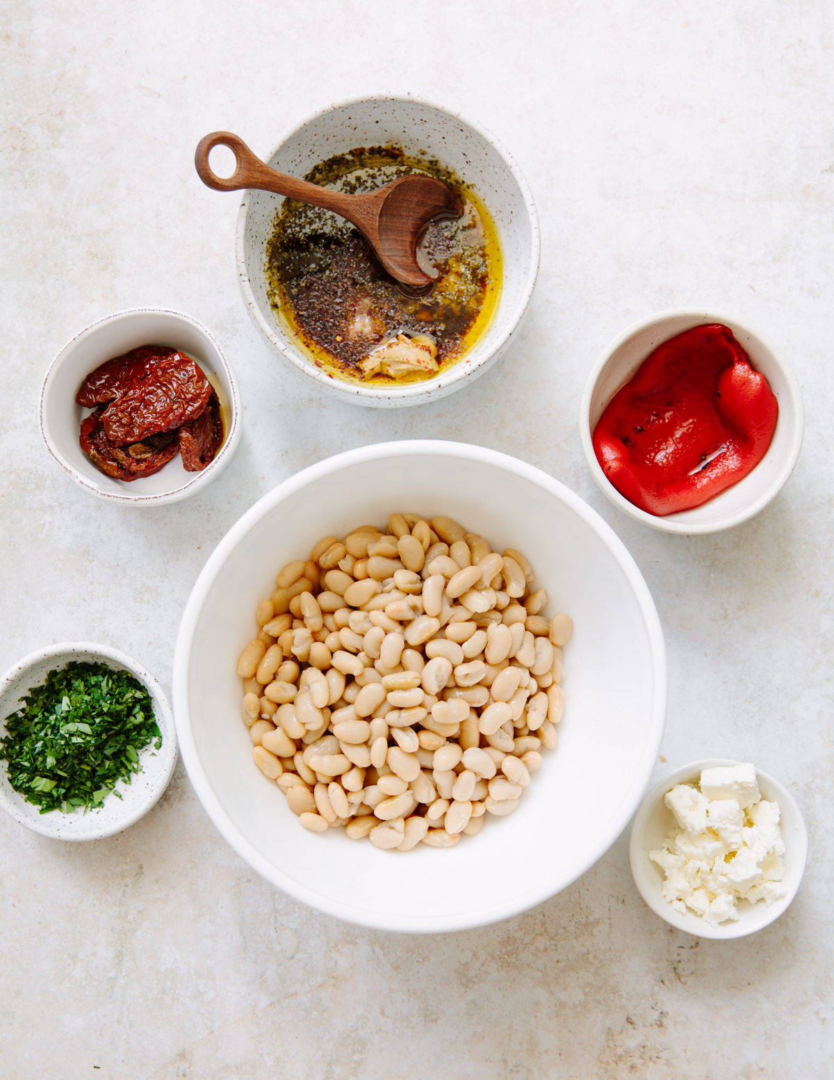 A top view of six bowls containing ingredients: white beans, chopped herbs, sun-dried tomatoes, feta cheese, roasted red peppers, and a bowl of olive oil with spices and a wooden spoon, all on a light surface.