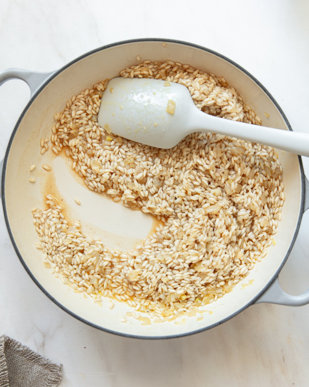 A white pot containing uncooked risotto rice being stirred with a white spatula; some rice is pushed aside, revealing the bottom of the pot.
