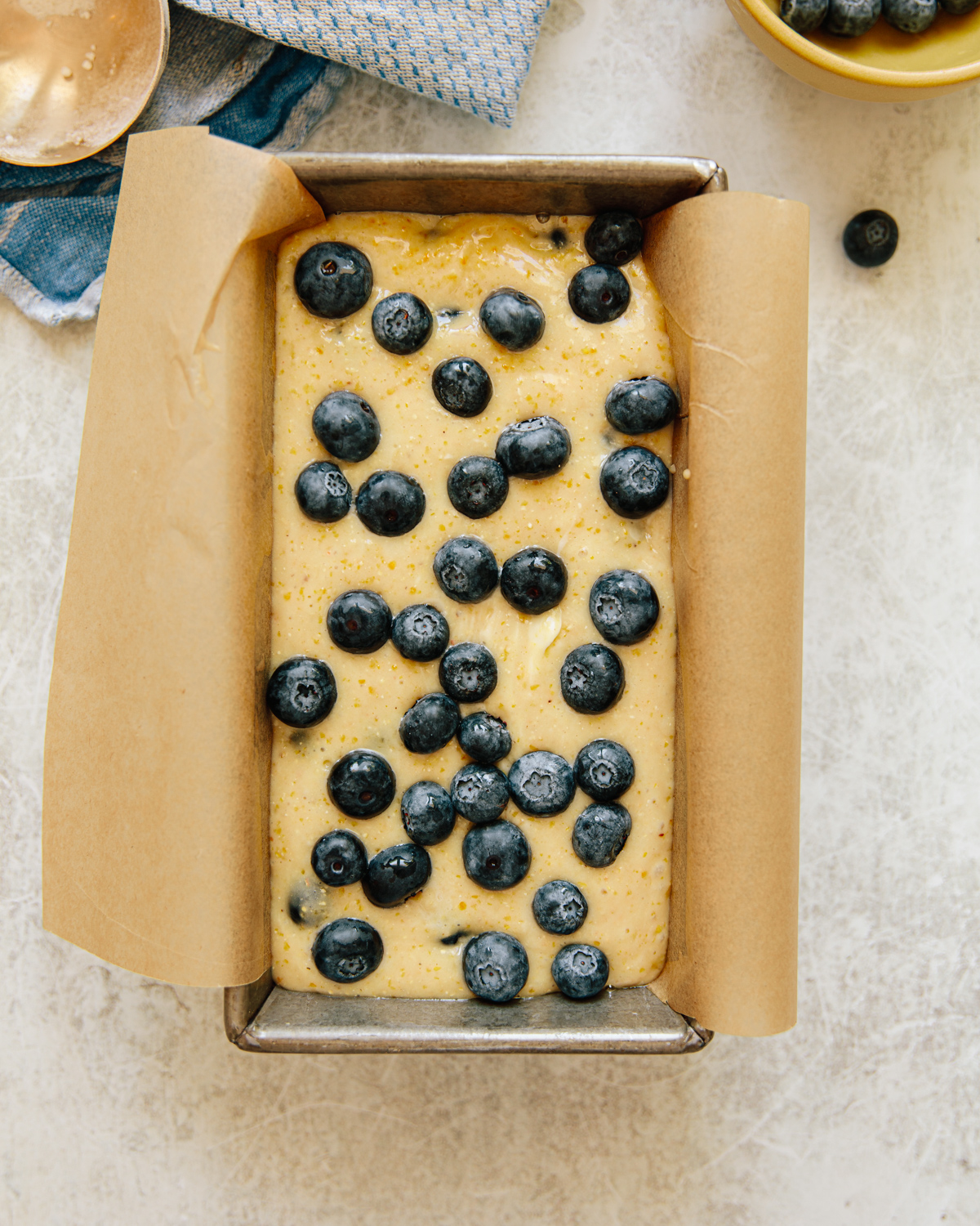 A loaf pan lined with parchment paper is filled with raw batter topped with fresh blueberries, ready to be baked. A blue cloth, a spoon, and a bowl of blueberries are nearby on a light surface.