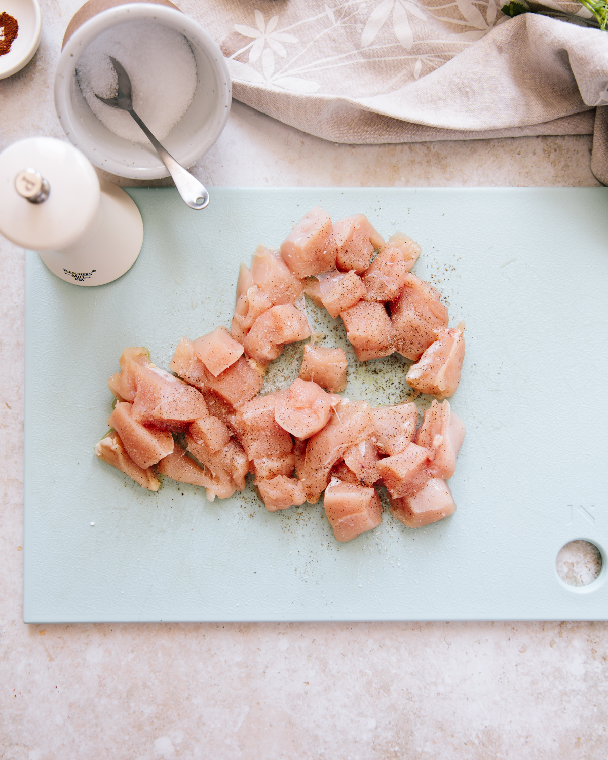 Raw, cubed chicken pieces are spread on a light blue cutting board, sprinkled with salt and pepper. A white bowl of salt with a spoon and a white pepper grinder are nearby on a light-colored countertop.
