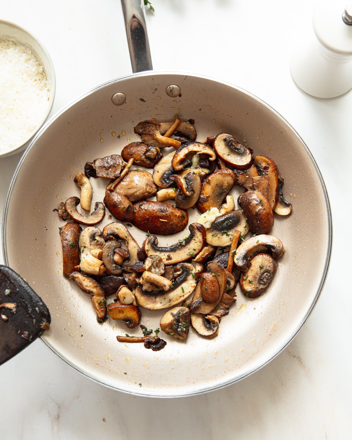 A pan filled with sautéed mushrooms sits on a white countertop, next to a bowl of grated cheese and a white pepper grinder.