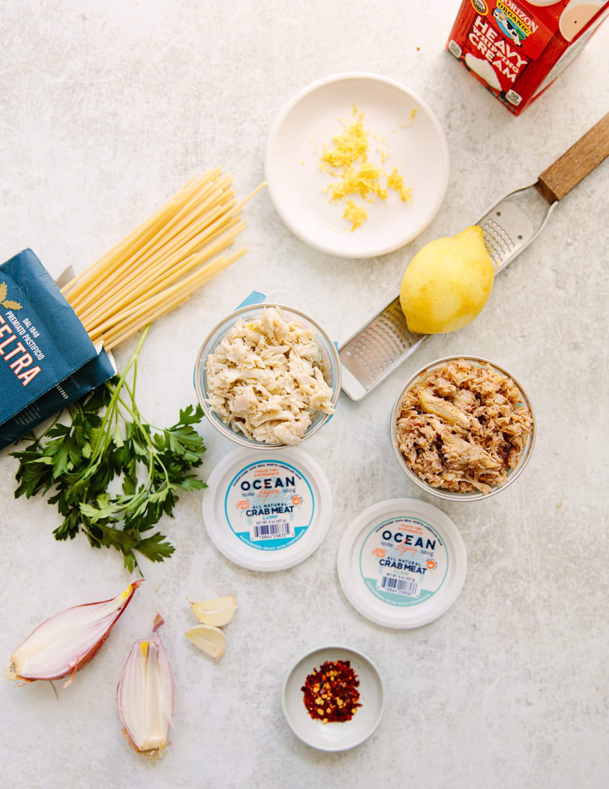 Overhead view of ingredients for a crab pasta dish, including packages of crab meat, spaghetti, parsley, shallots, garlic, lemon, lemon zest, red pepper flakes, and heavy cream, arranged on a white surface.