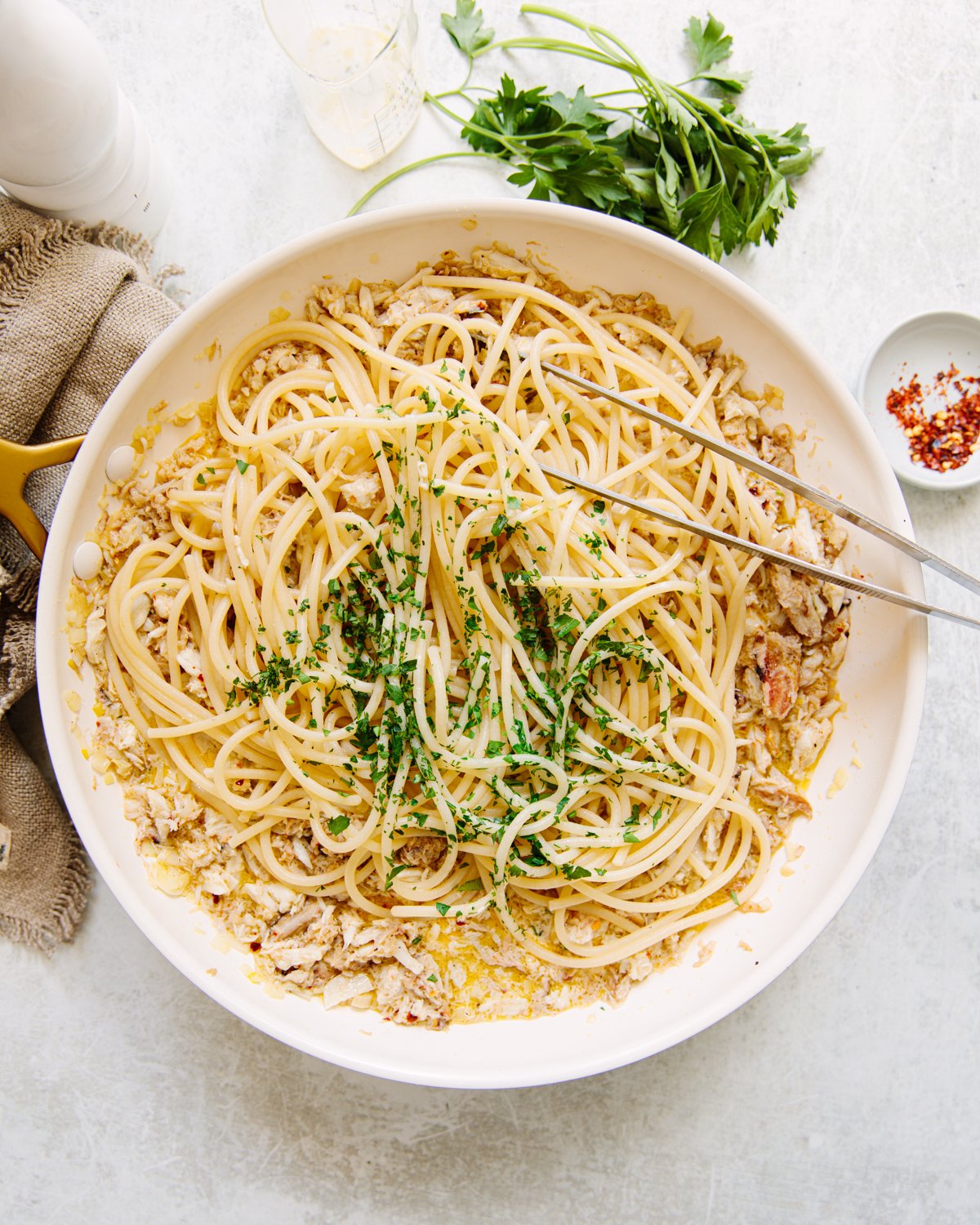 A white skillet filled with cooked spaghetti, crab, creamy sauce, garnished with chopped parsley. Metal tongs rest on the pasta. Fresh parsley, a napkin, and a small dish of chili flakes are nearby.