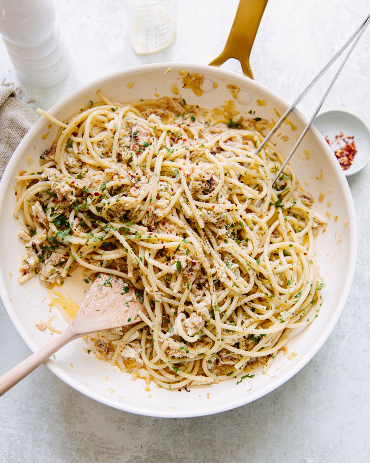 A pan of cooked spaghetti mixed with a creamy crab meat sauce, garnished with chopped parsley. A wooden spoon and serving fork rest in the pan, and a small dish of red pepper flakes sits nearby.