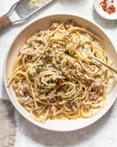 A bowl of spaghetti topped with shredded crab meat, chopped herbs, and red pepper flakes, with a fork twirled in the pasta. A grater with lemon zest and a small dish of chili flakes are nearby.