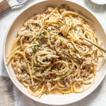 A bowl of spaghetti topped with shredded crab meat, chopped herbs, and red pepper flakes, with a fork twirled in the pasta. A grater with lemon zest and a small dish of chili flakes are nearby.