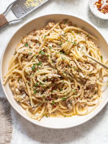 A bowl of spaghetti topped with shredded crab meat, chopped herbs, and red pepper flakes, with a fork twirled in the pasta. A grater with lemon zest and a small dish of chili flakes are nearby.