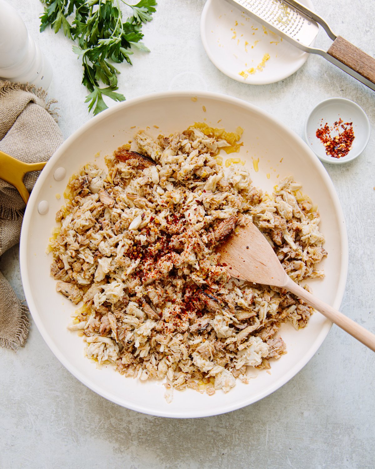 A skillet filled with crabmeat, sprinkled with red pepper flakes, sits on a light countertop. A wooden spatula rests in the pan. Parsley sprigs, a grater, and small dishes with spices are nearby.