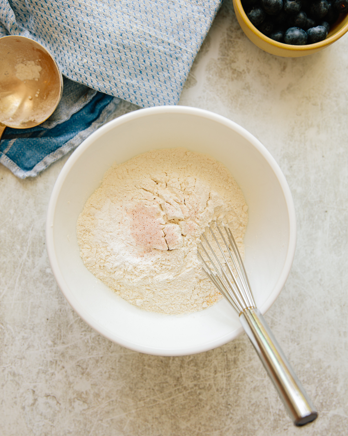 A white bowl with flour mixture and a metal whisk on a light countertop, next to a blue-striped towel, a gold measuring cup, and a small yellow bowl of blueberries.
