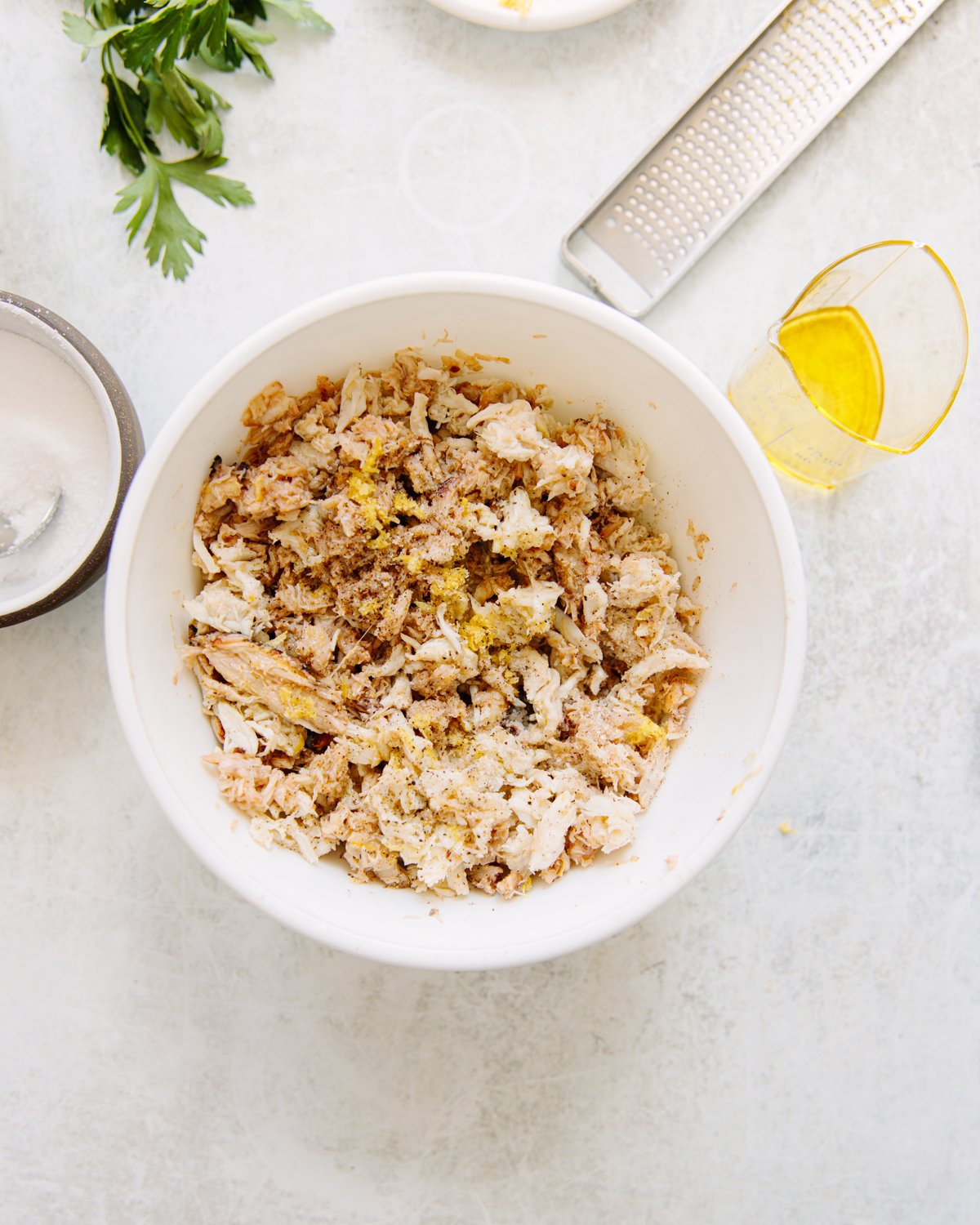 A white bowl filled with shredded chicken mixed with seasoning sits on a light surface. Nearby are a glass of olive oil, a grater, parsley leaves, and a bowl of flour.