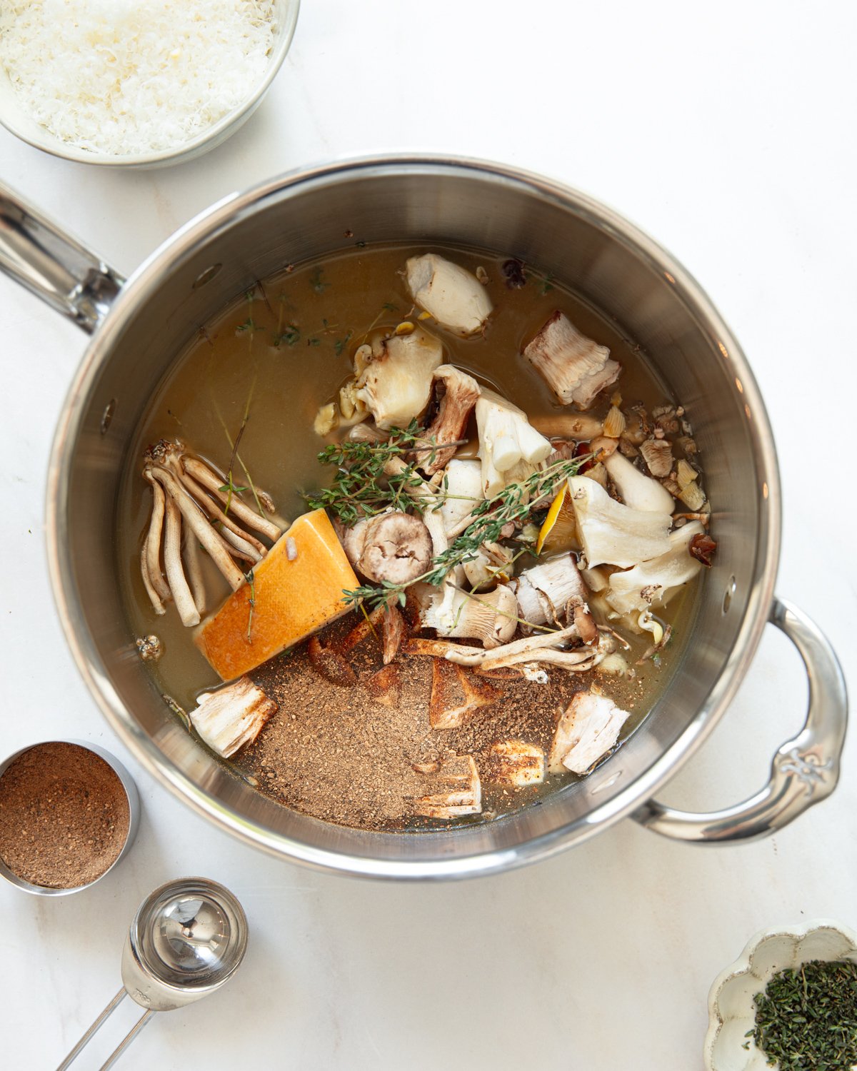 A stainless steel pot filled with mushroom trimmings, fresh herbs, a Parmesan cheese rind, and broth is surrounded by measuring spoons and small bowls on a white countertop.
