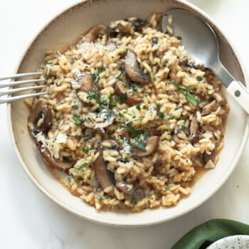 A bowl of creamy mushroom risotto garnished with chopped herbs and grated cheese, served with a spoon and fork on a light-colored surface.