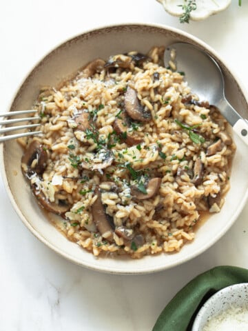 A bowl of creamy mushroom risotto garnished with chopped herbs and grated cheese, served with a spoon and fork on a light-colored surface.