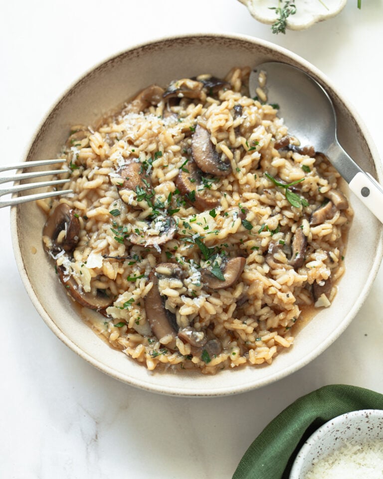 A bowl of creamy mushroom risotto garnished with chopped herbs and grated cheese, served with a spoon and fork on a light-colored surface.