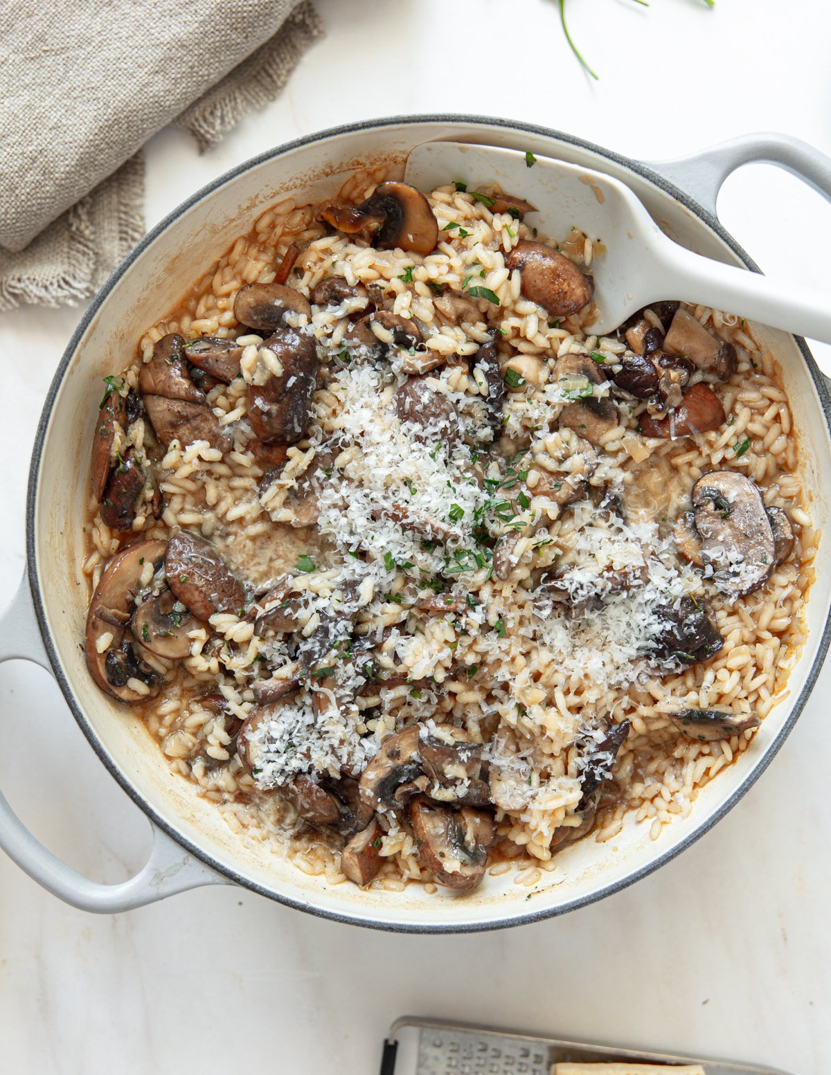 A pot of creamy mushroom risotto topped with grated cheese and herbs sits on a white surface, with a beige cloth and a cheese grater partially visible beside it.
