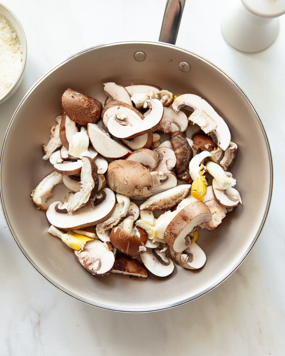 A frying pan filled with sliced mushrooms ready to be cooked, on a white countertop next to a bowl of grated cheese and a white pepper grinder.