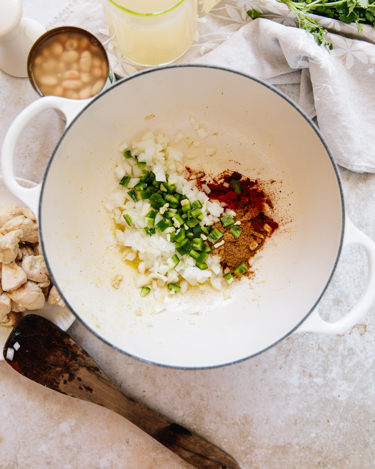 A white pot contains chopped onions, green peppers, and several ground spices. A wooden spoon rests nearby, with beans, broth, herbs, and chopped chicken arranged around the pot on a light countertop.