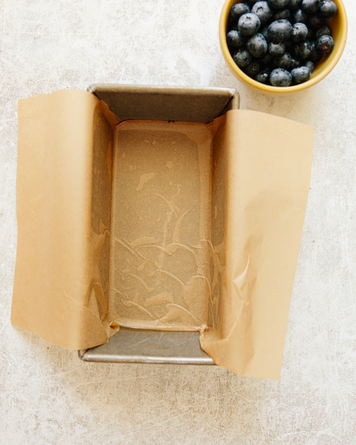 A loaf pan lined with parchment paper sits on a light surface next to a small yellow bowl filled with fresh blueberries.