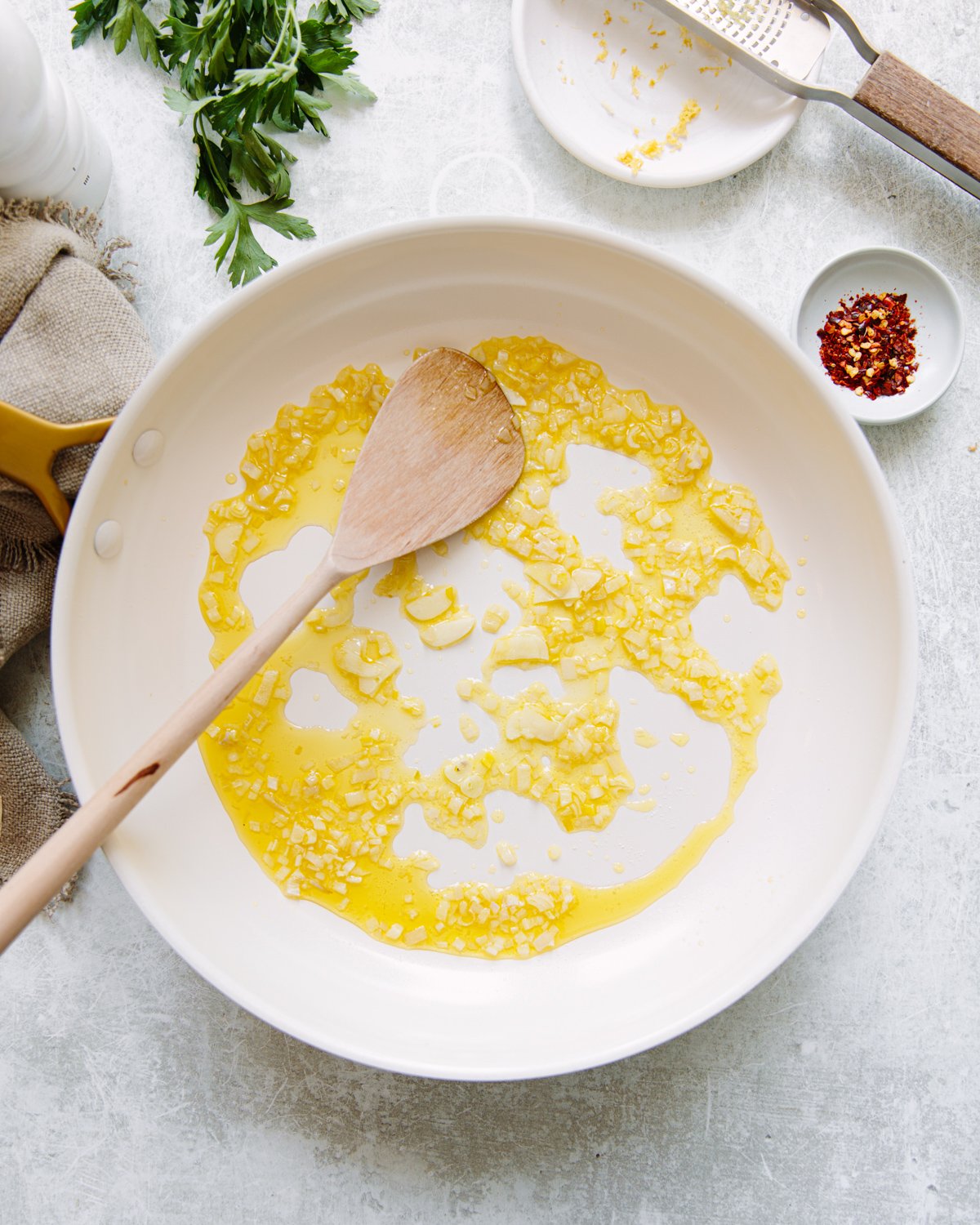 A white skillet with chopped garlic sautéing in olive oil, stirred with a wooden spoon. Fresh parsley, a grater, chili flakes, and a kitchen towel are on the light countertop nearby.