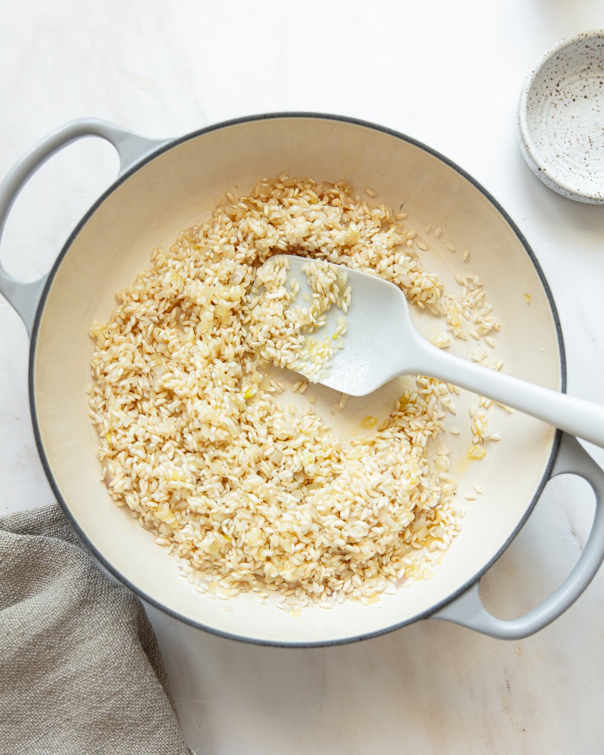 A white pot with handles contains uncooked rice being stirred with a white spatula. A small bowl and a beige cloth are nearby on a white countertop.