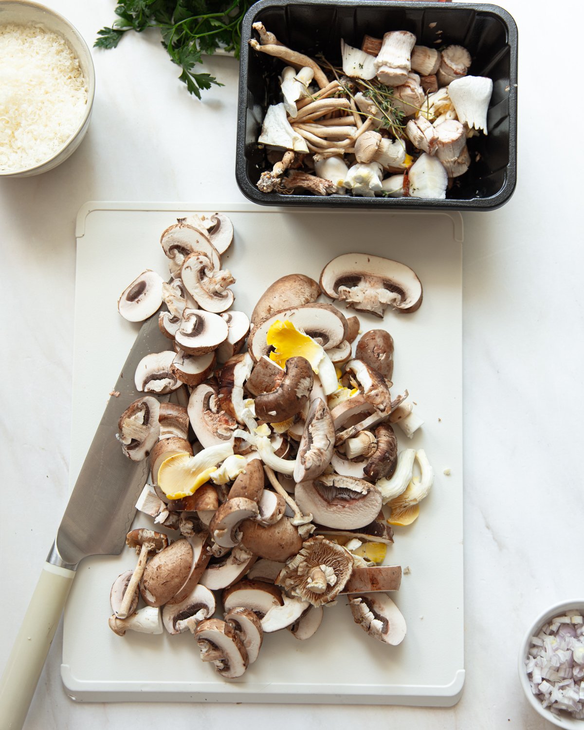 A cutting board with sliced mushrooms and a knife, next to a black tray of whole and trimmed mushrooms. Bowls with grated cheese and chopped shallots are nearby on a white surface.