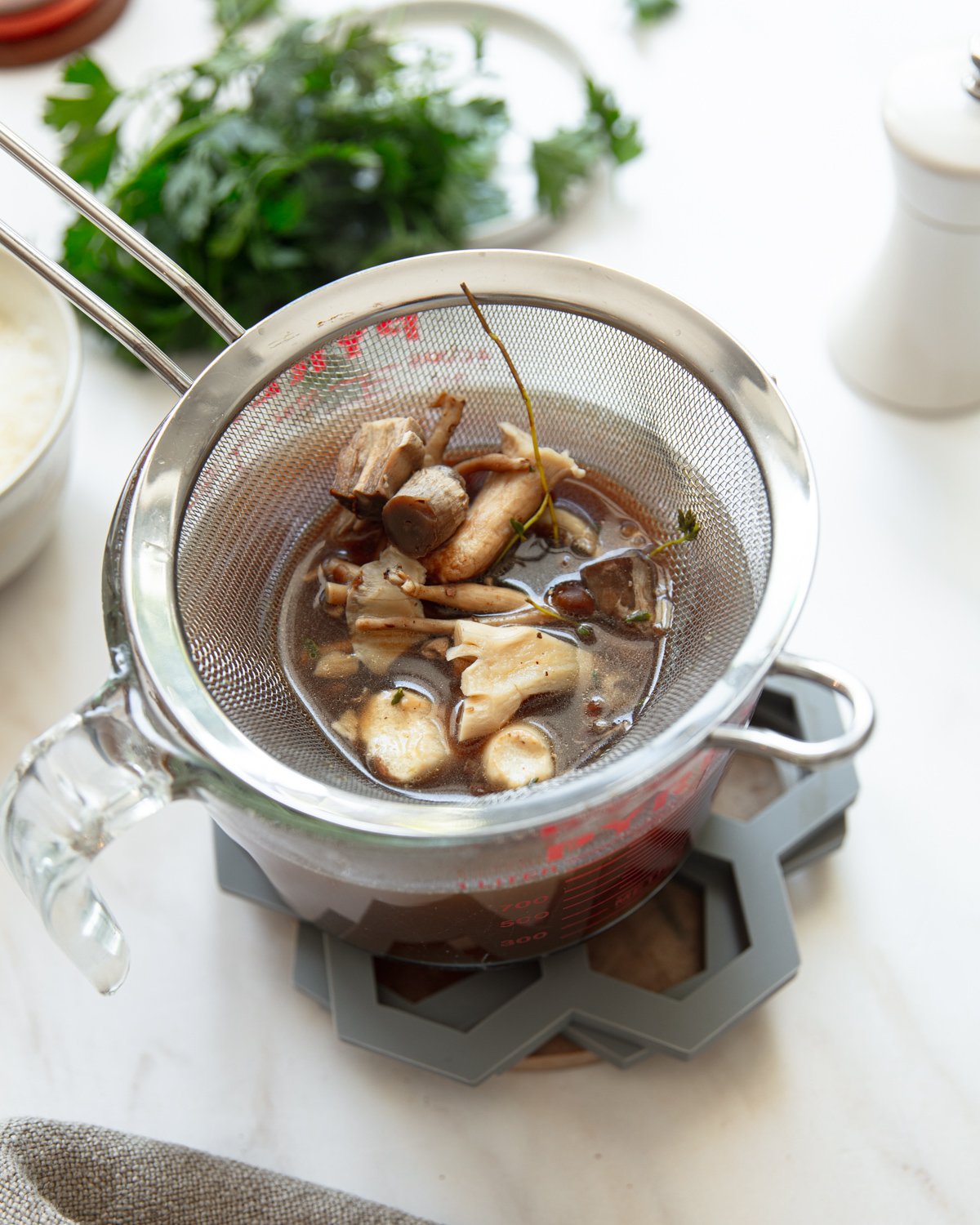 A metal strainer with cooked mushrooms and herbs rests over a glass measuring cup filled with dark broth. Fresh herbs, a bowl of rice, and a pepper grinder are visible in the background on a white surface.
