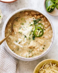 A bowl of creamy white chili with white beans, shredded cheese, sliced jalapeños, and herbs, with a gold spoon in the bowl and garnishes of cheese and jalapeños nearby.