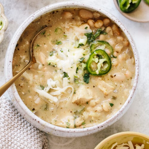 A bowl of creamy white chili with white beans, shredded cheese, sliced jalapeños, and herbs, with a gold spoon in the bowl and garnishes of cheese and jalapeños nearby.