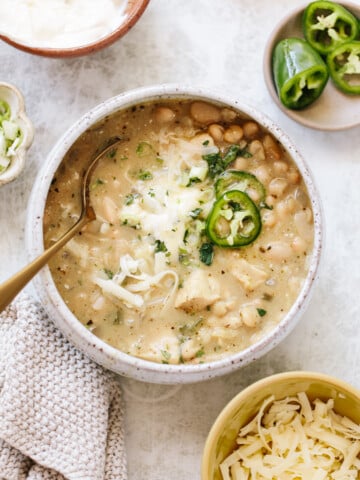 A bowl of creamy white chili topped with shredded cheese, sliced jalapeños, and herbs. Surrounding the bowl are dishes with sour cream, sliced green onions, jalapeño slices, and extra shredded cheese. A spoon rests in the chili.