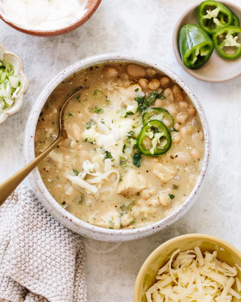 A bowl of creamy white chili topped with shredded cheese, sliced jalapeños, and herbs. Surrounding the bowl are dishes with sour cream, sliced green onions, jalapeño slices, and extra shredded cheese. A spoon rests in the chili.