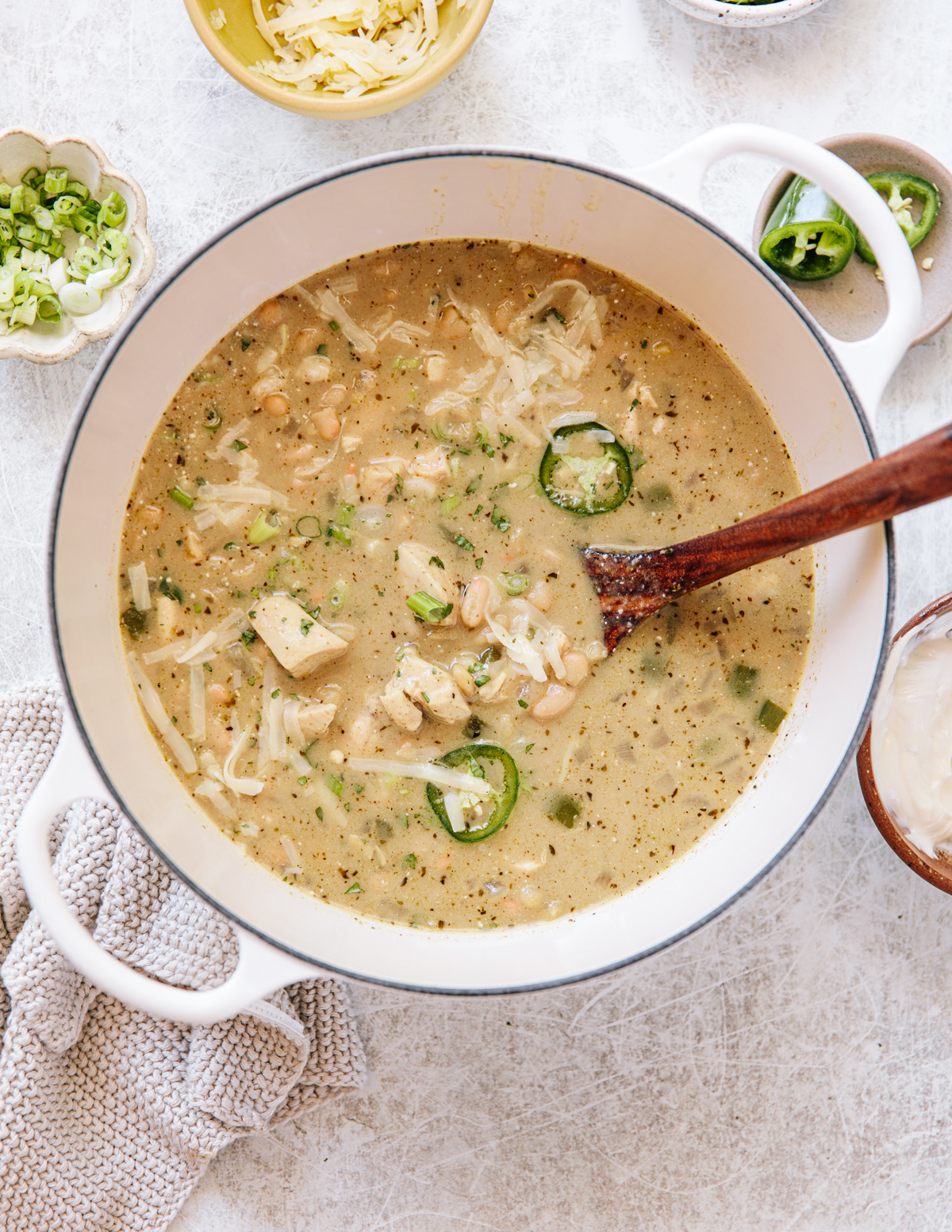 A pot of creamy white chili with chicken, white beans, shredded cheese, sliced jalapeños, and chopped green onions, with a wooden spoon. Small bowls of cheese, scallions, and jalapeños are nearby on a light surface.