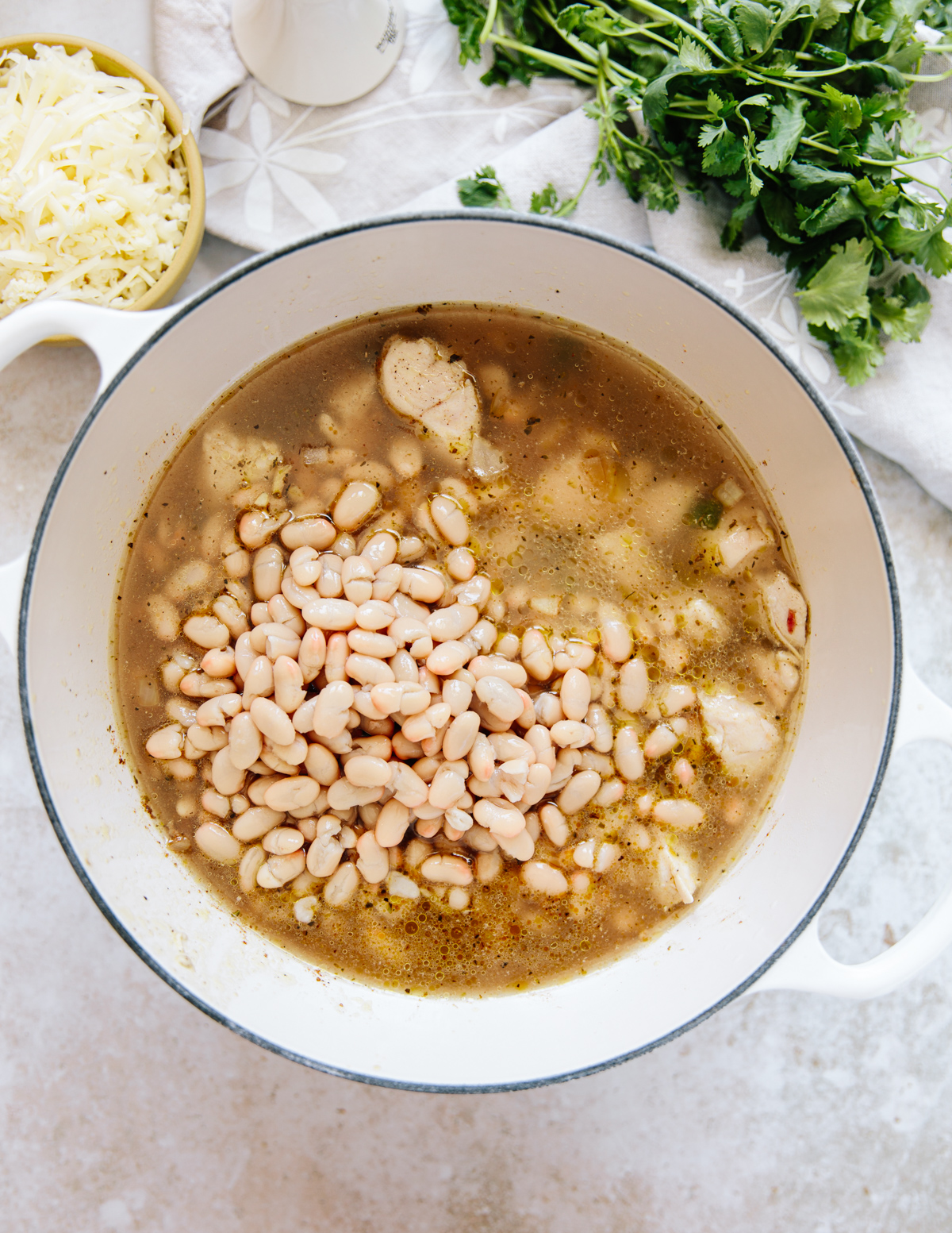 A white pot filled with broth, cooked chicken pieces, and a heap of white beans. Nearby are a bowl of shredded cheese and a bunch of fresh cilantro on a light countertop.