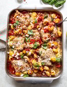 A baking dish filled with roasted chicken thighs, golden gnocchi, cherry tomatoes, fresh basil leaves, and crumbled goat cheese, all baked together and served on a light-colored surface.