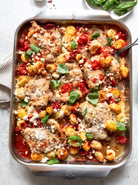 A baking dish filled with roasted chicken thighs, golden gnocchi, cherry tomatoes, fresh basil leaves, and crumbled goat cheese, all baked together and served on a light-colored surface.