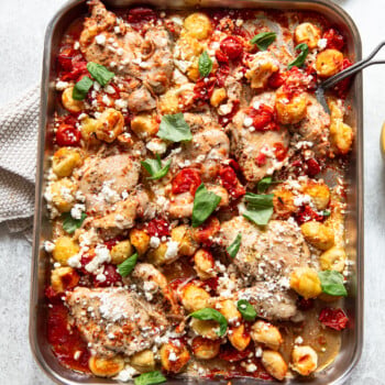 A baking dish filled with roasted chicken thighs, golden gnocchi, cherry tomatoes, fresh basil leaves, and crumbled goat cheese, all baked together and served on a light-colored surface.