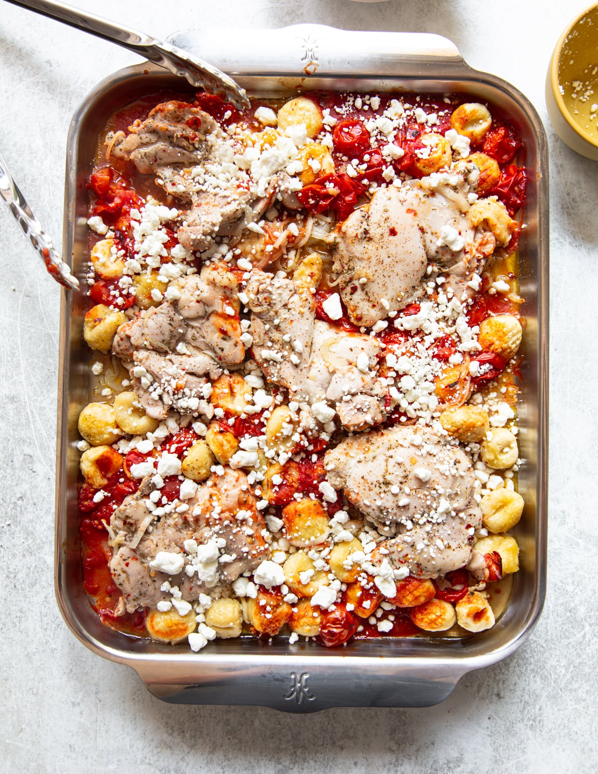 A sheet pan filled with seasoned chicken thighs, roasted cherry tomatoes, gnocchi, and crumbled goat cheese, with metal tongs resting on the edge of the dish.