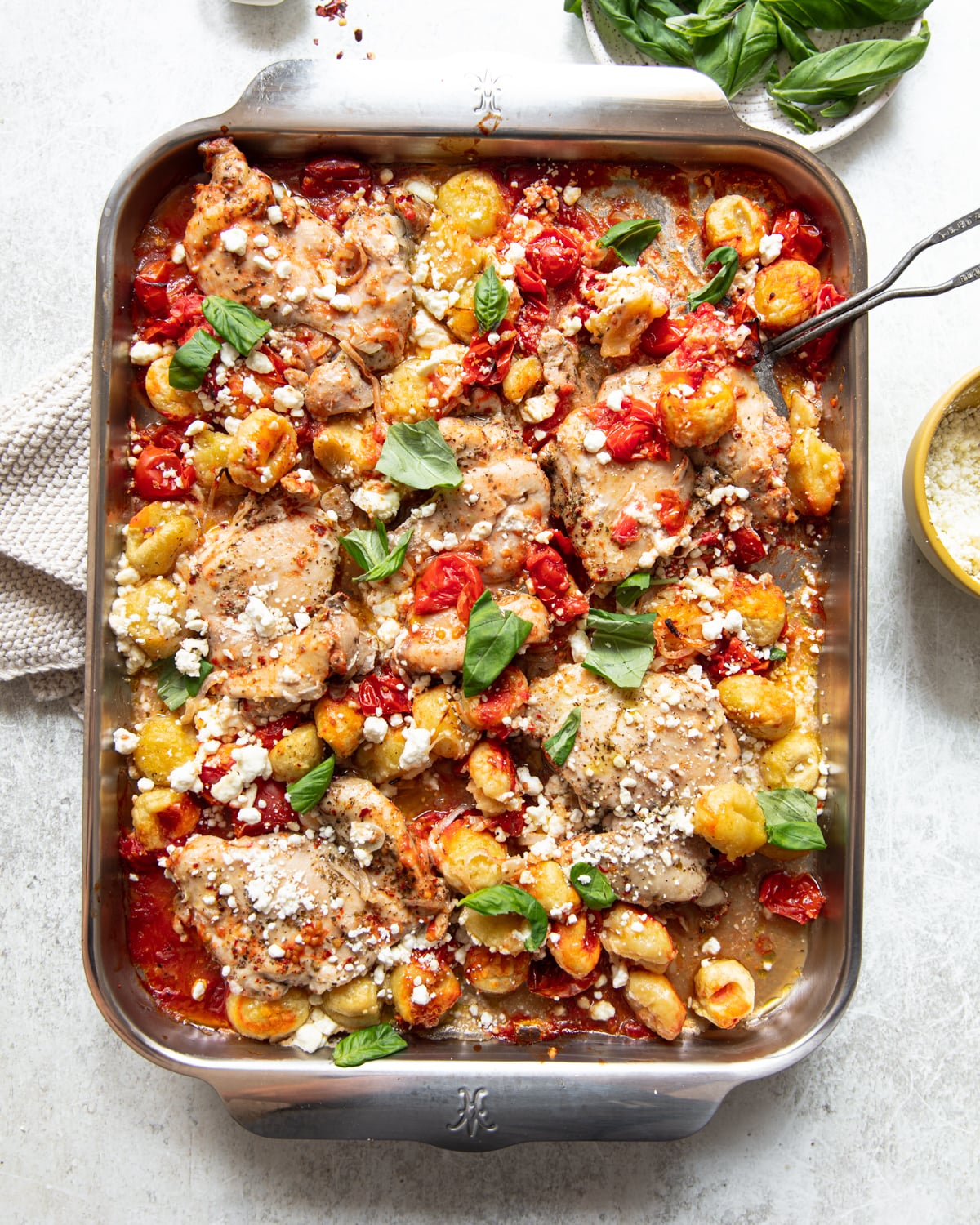 A baking dish filled with baked chicken thighs, cherry tomatoes, gnocchi, fresh basil, and crumbled cheese, with a serving spoon, a bowl of grated cheese, and a kitchen towel nearby.