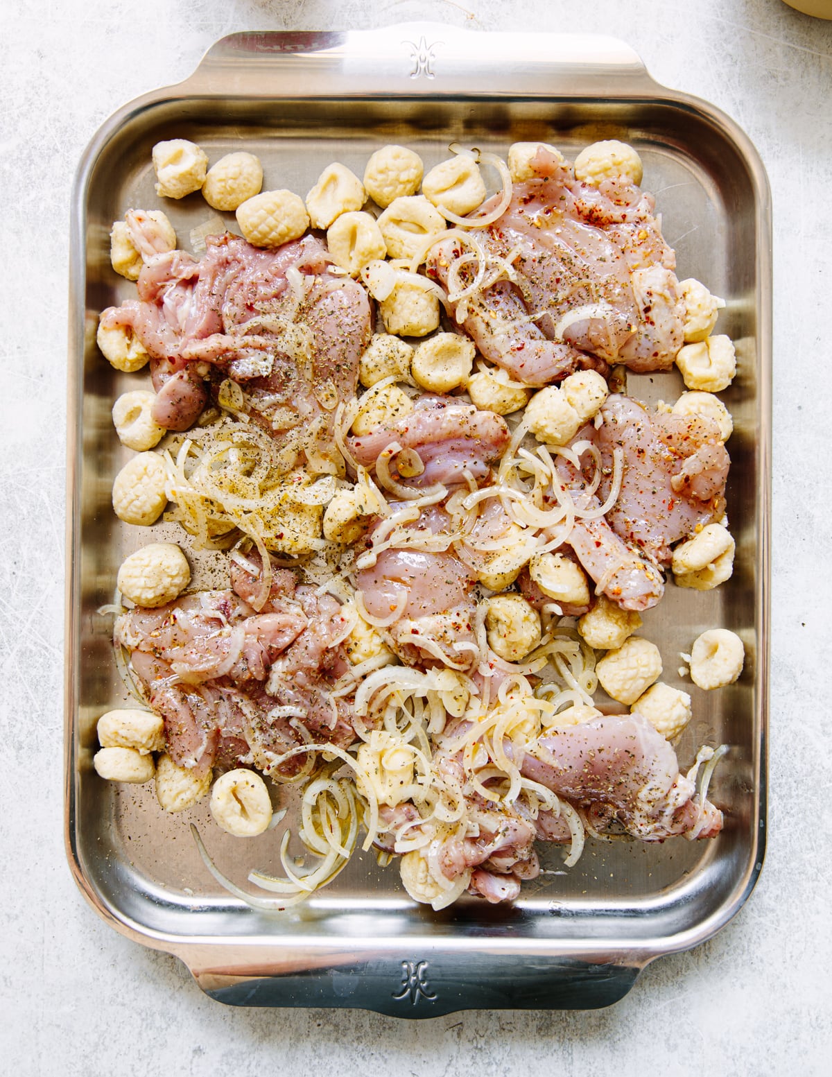 Raw seasoned chicken thighs, sliced onions, and uncooked gnocchi are spread out on a large stainless steel baking tray, ready to be cooked.
