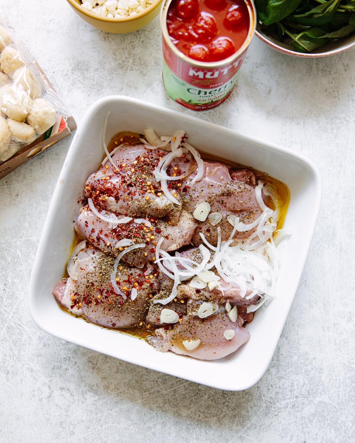 Raw chicken thighs in a white dish, topped with sliced onions, garlic, and spices. Surrounding the dish are a can of cherry tomatoes, a bowl of fresh basil, crumbled goat cheese, and a bag of gnocchi.