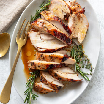 Sliced roasted turkey breast garnished with fresh rosemary and thyme on a white oval plate, with a gold fork and spoon beside a beige napkin. Juices from the turkey are visible on the plate.