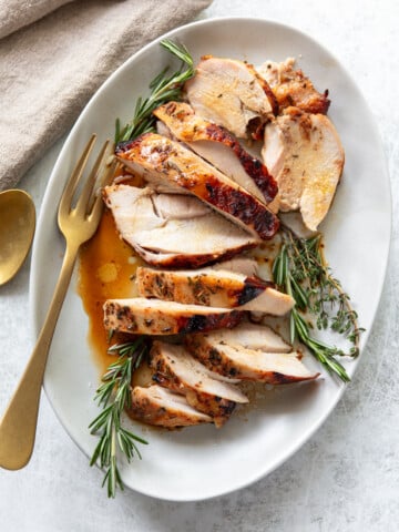 Sliced roasted turkey breast garnished with fresh rosemary and thyme on a white oval plate, with a gold fork and spoon beside a beige napkin. Juices from the turkey are visible on the plate.