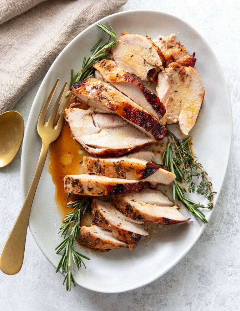 Sliced roasted turkey breast garnished with fresh rosemary and thyme on a white oval plate, with a gold fork and spoon beside a beige napkin. Juices from the turkey are visible on the plate.