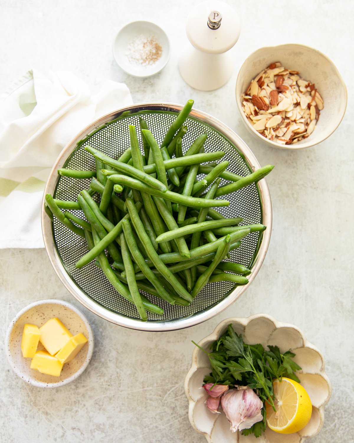 Overhead view of fresh green beans in a colander, surrounded by small bowls containing sliced almonds, butter, garlic, parsley, half a lemon, and a small dish of salt, on a light-colored surface.
