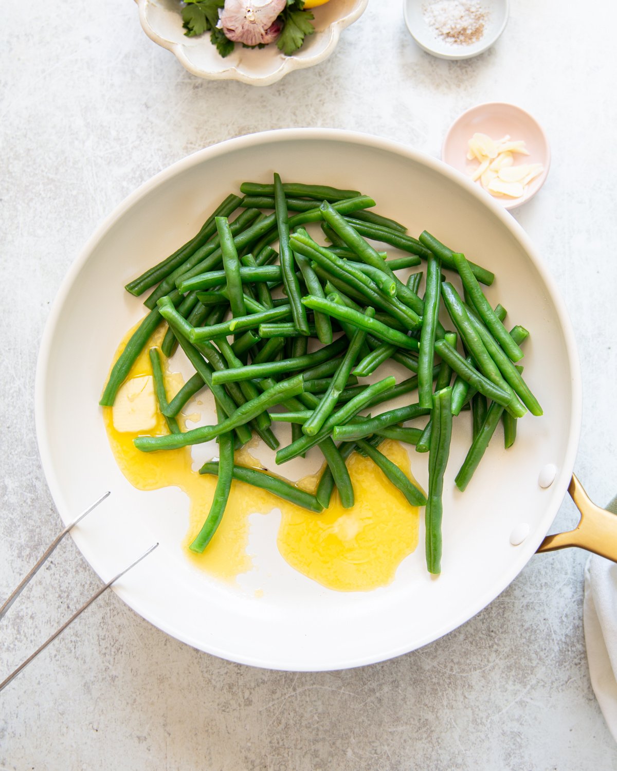 A white skillet with cooked green beans and melted butter, placed on a light-colored surface with small dishes containing garlic, salt, and herbs nearby.
