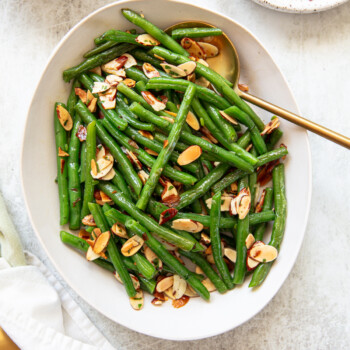An oval white platter filled with green bean almondine, topped with toasted almond slices, garnished with chopped parsley. A gold serving spoon rests in the dish, and a small plate with extra almond slices is nearby.