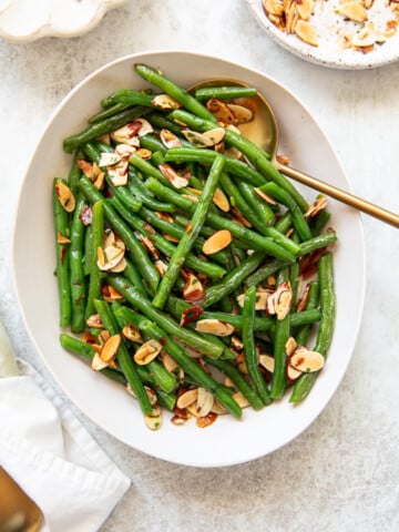 An oval white platter filled with green bean almondine, topped with toasted almond slices, garnished with chopped parsley. A gold serving spoon rests in the dish, and a small plate with extra almond slices is nearby.
