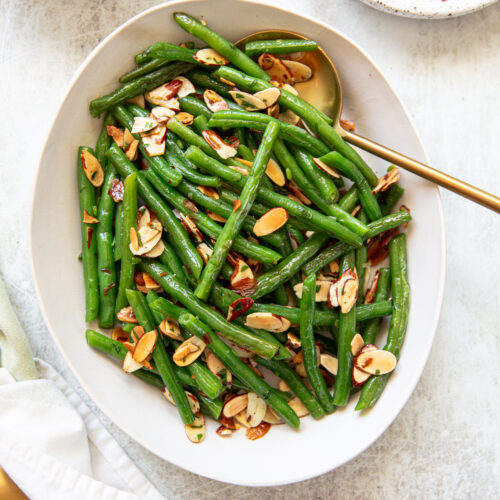 An oval white platter filled with green bean almondine, topped with toasted almond slices, garnished with chopped parsley. A gold serving spoon rests in the dish, and a small plate with extra almond slices is nearby.