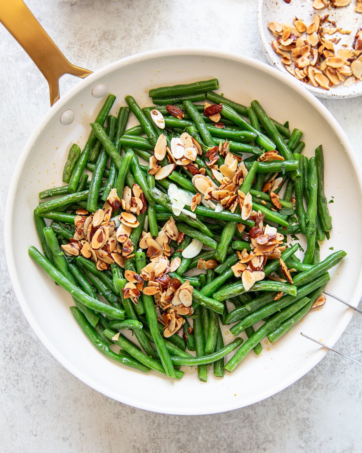 A white skillet filled with sautéed green beans topped with toasted sliced almonds sits on a light countertop, with a plate of extra almonds nearby.