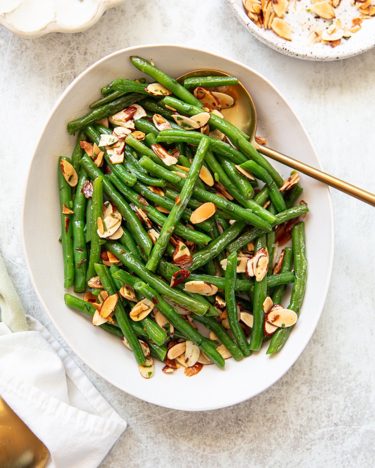 An oval white platter filled with green bean almondine, topped with toasted almond slices, garnished with chopped parsley. A gold serving spoon rests in the dish, and a small plate with extra almond slices is nearby.